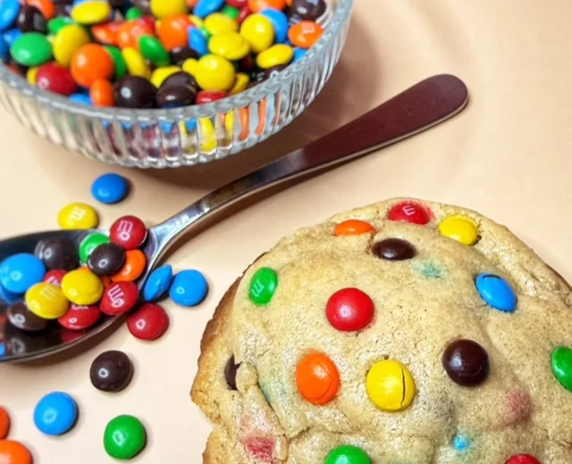 The Happy Chapter cookie studded with colorful M&M’s next to a bowl and spoonful of chocolate candies