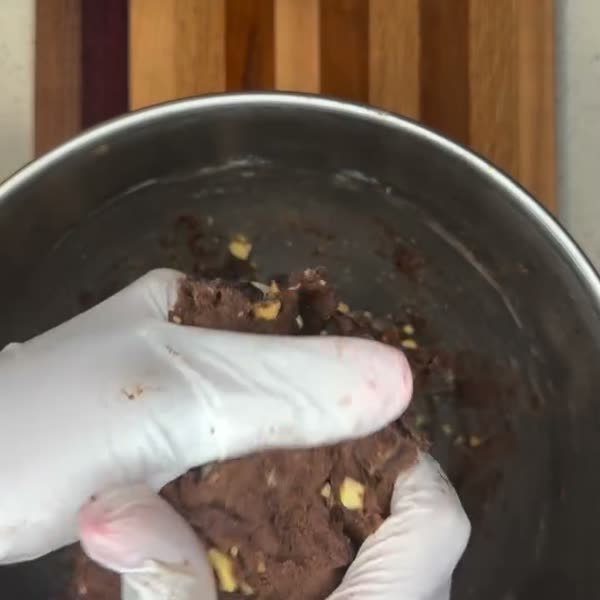 Gloved hands folding chocolate cookie dough by hand in a steel bowl